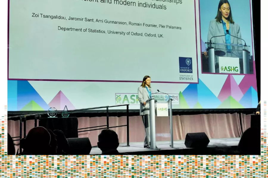 A photo of Zoi standing at a podium giving her plenary talk. She is a white woman wearing a grey suit and a light blue collared shirt. The presentation screen is behind her with a close-up of her head and shoulders next to her presentation slides. The photo is against a white background with a green, orange, and light blue graphical representation of genomes at the bottom.