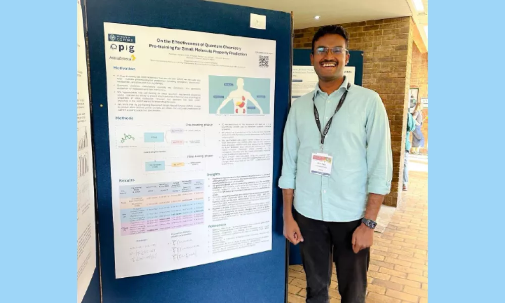 A photo of a tall brown man with glasses standing to the left of a poster featuring his research. He wears a light blue shirt and black trousers with light tan coloured shoes.