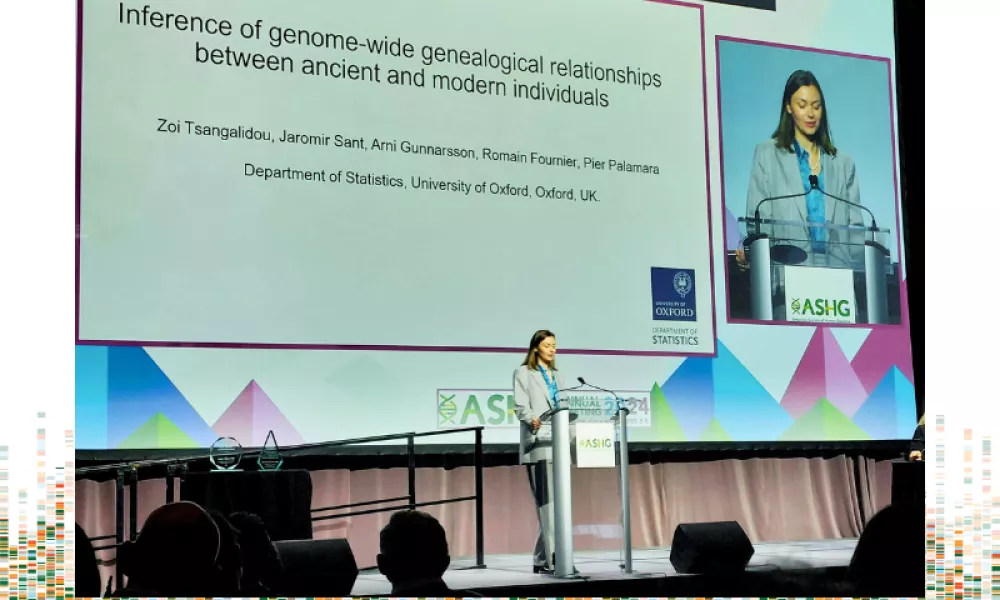 A photo of Zoi standing at a podium giving her plenary talk. She is a white woman wearing a grey suit and a light blue collared shirt. The presentation screen is behind her with a close-up of her head and shoulders next to her presentation slides. The photo is against a white background with a green, orange, and light blue graphical representation of genomes at the bottom.